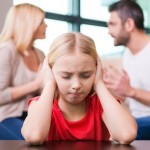 Please stop fighting! Depressed little girl leaning at the table and covering ear with hands while her parents shouting at each other in the background